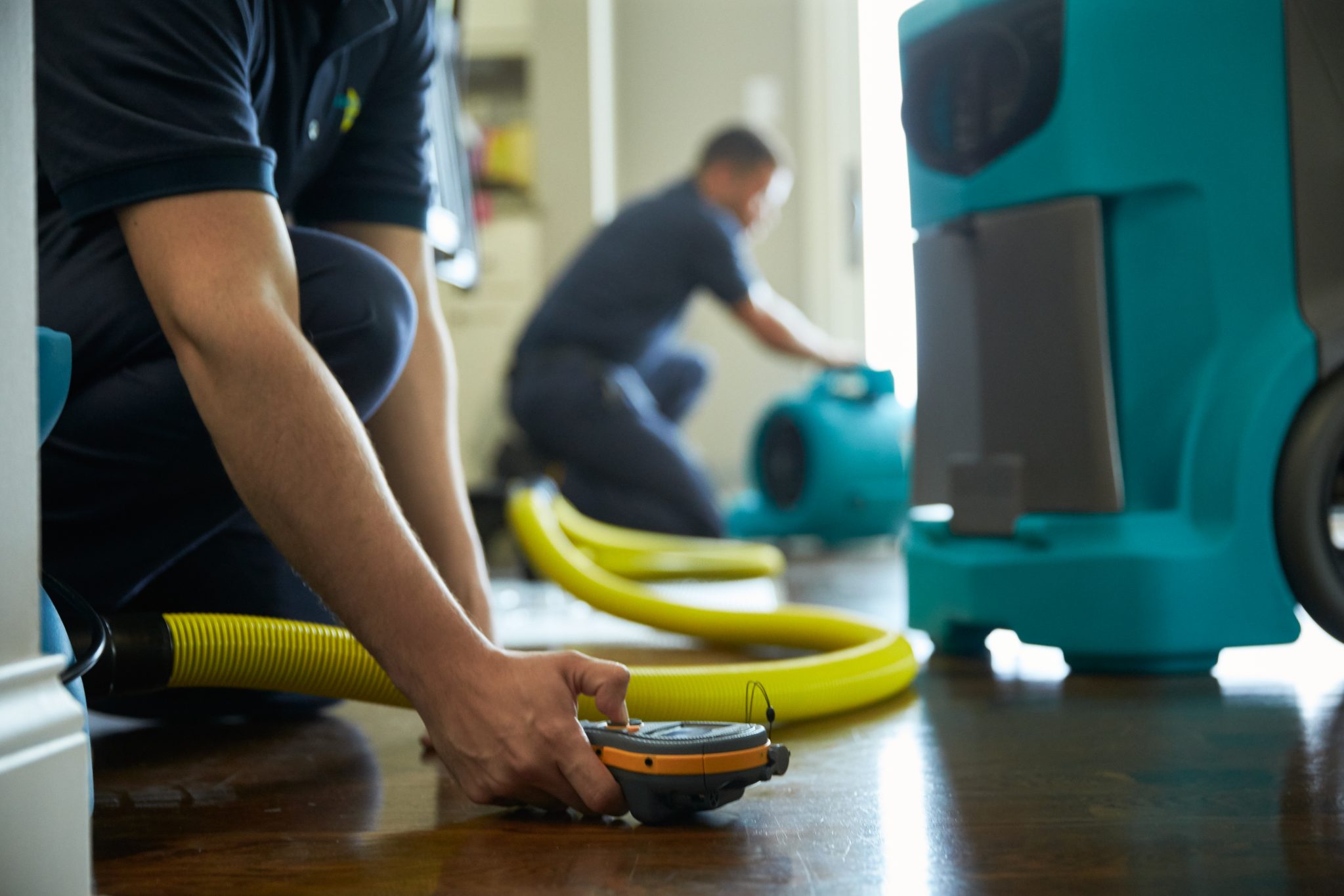 Close-up of a water damage restoration technician setting up equipment with hoses and moisture meters, while another technician operates an air mover in the background. This image illustrates NSH Home Services' professional approach to water damage recovery, using advanced tools to ensure thorough drying and restoration.