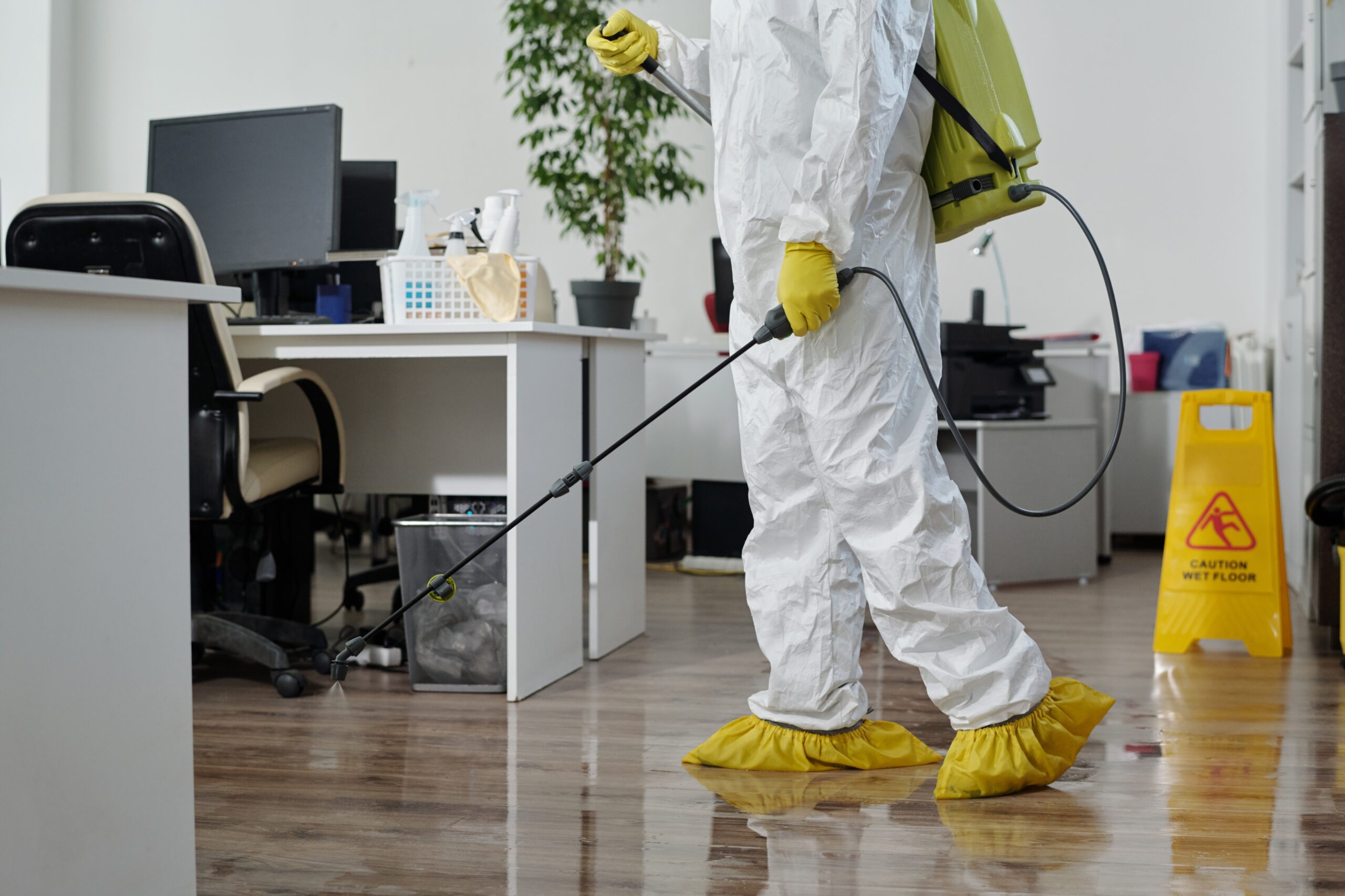 person wearing a full protective suit, including a face mask, gloves, and a hood, engaged in a pest control or disinfection activity inside a home. They are holding a sprayer with a long nozzle, likely used for applying chemicals or disinfectants, while a red tank on their back holds the solution. The setting appears to be a residential space with natural light coming through a window, highlighting the clean, controlled environment