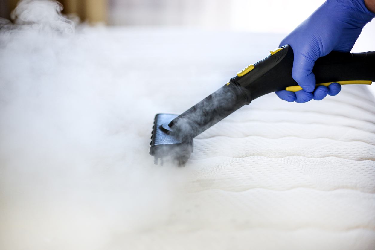 Professional mattress steam cleaning service using high-temperature steam to sanitize and remove deep-seated allergens. Technician wearing protective gloves for a thorough and hygienic process, provided by NSH Home Services.