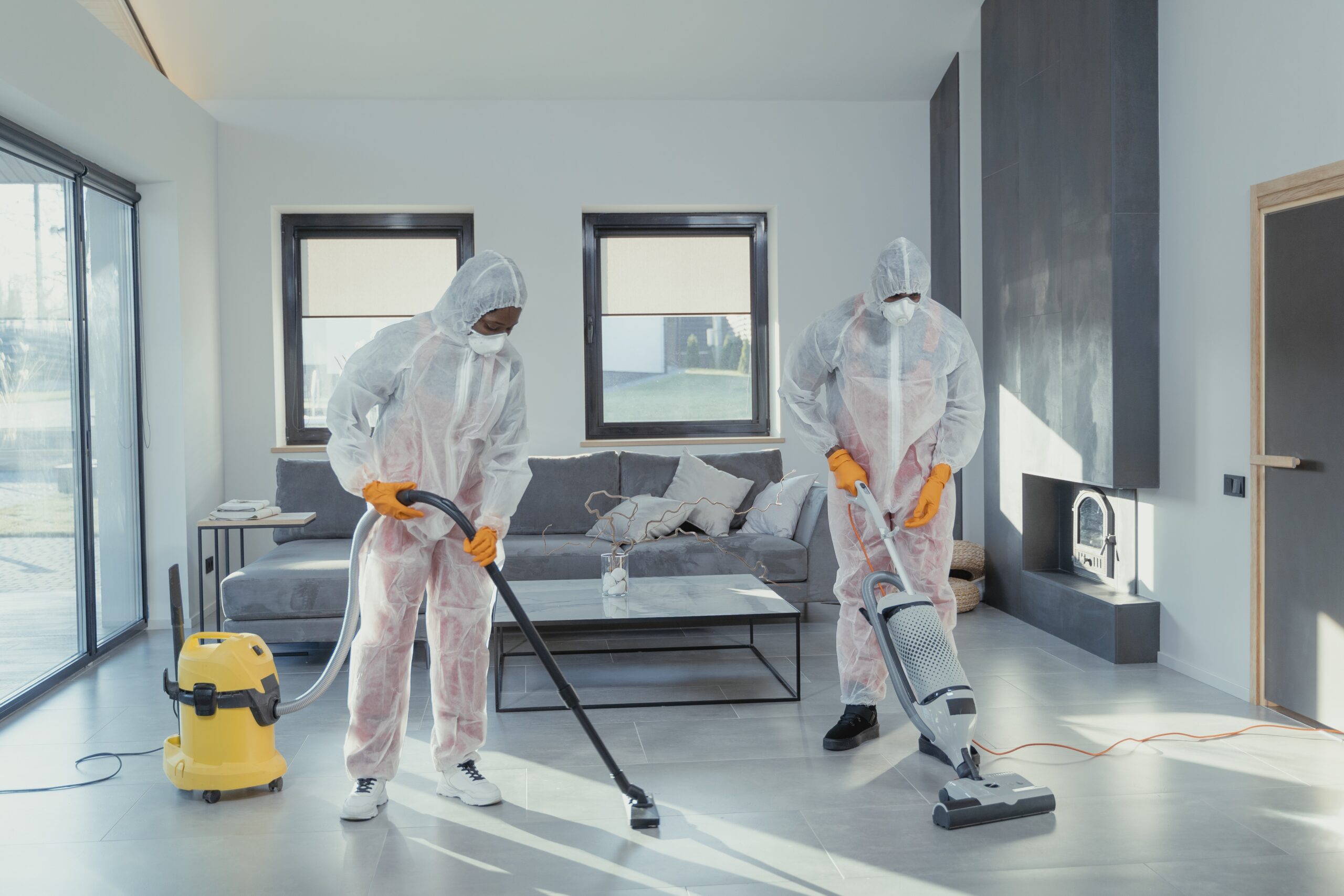 Two technicians in protective suits performing a comprehensive deep cleaning in a modern living room. Equipped with professional-grade vacuum and cleaning tools, ensuring a sanitized and spotless environment, provided by NSH Home Services.