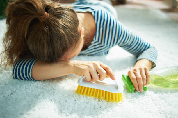 Closeup on a tired woman in a striped t-shirt and white pants with a spray bottle of green cleaning supplies and yellow brush laying on the white carpet at home on sunny day.